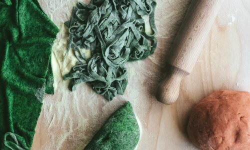 Green and pink hand-rolled tagliatelle beside a wooden rolling pin and coloured pasta dough